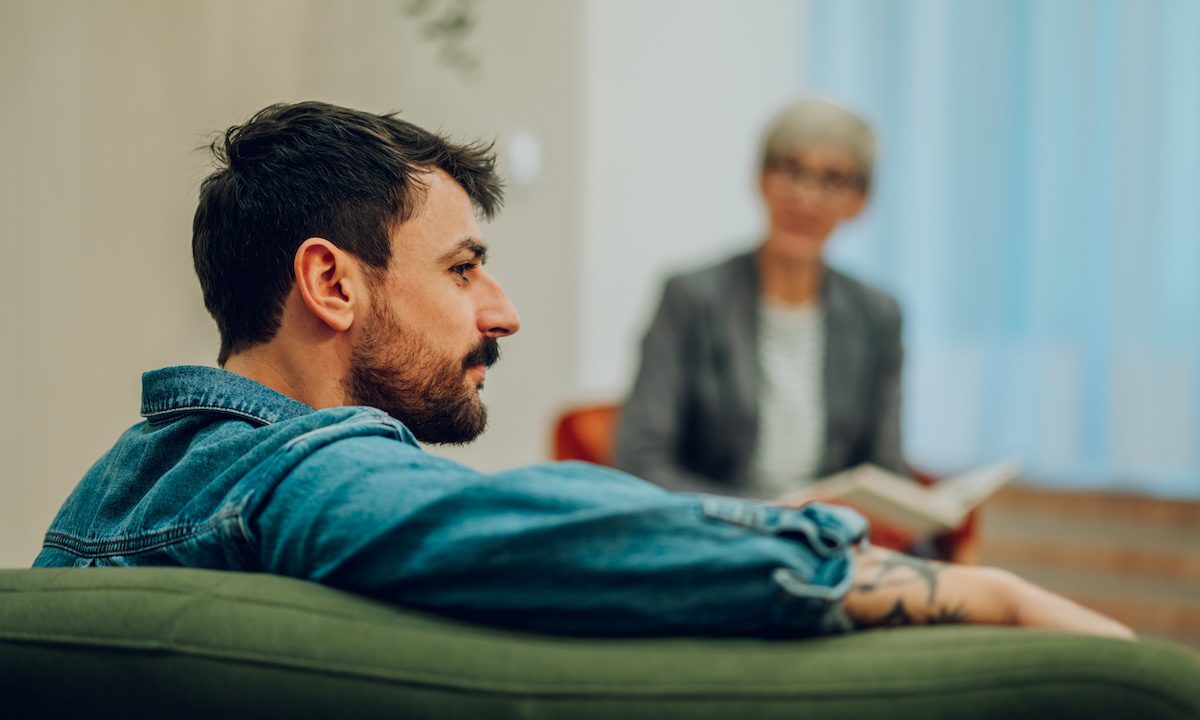 Profile of a young man with issues sitting on a sofa in the office with a psychotherapist and discussing trauma and problems. In a blurry background is the therapist giving support and giving advice.