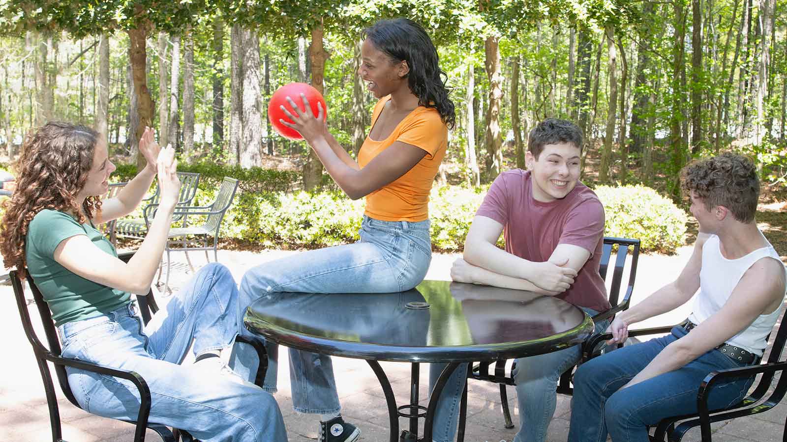 Four teenagers sitting around a table passing a red ball around and laughing.