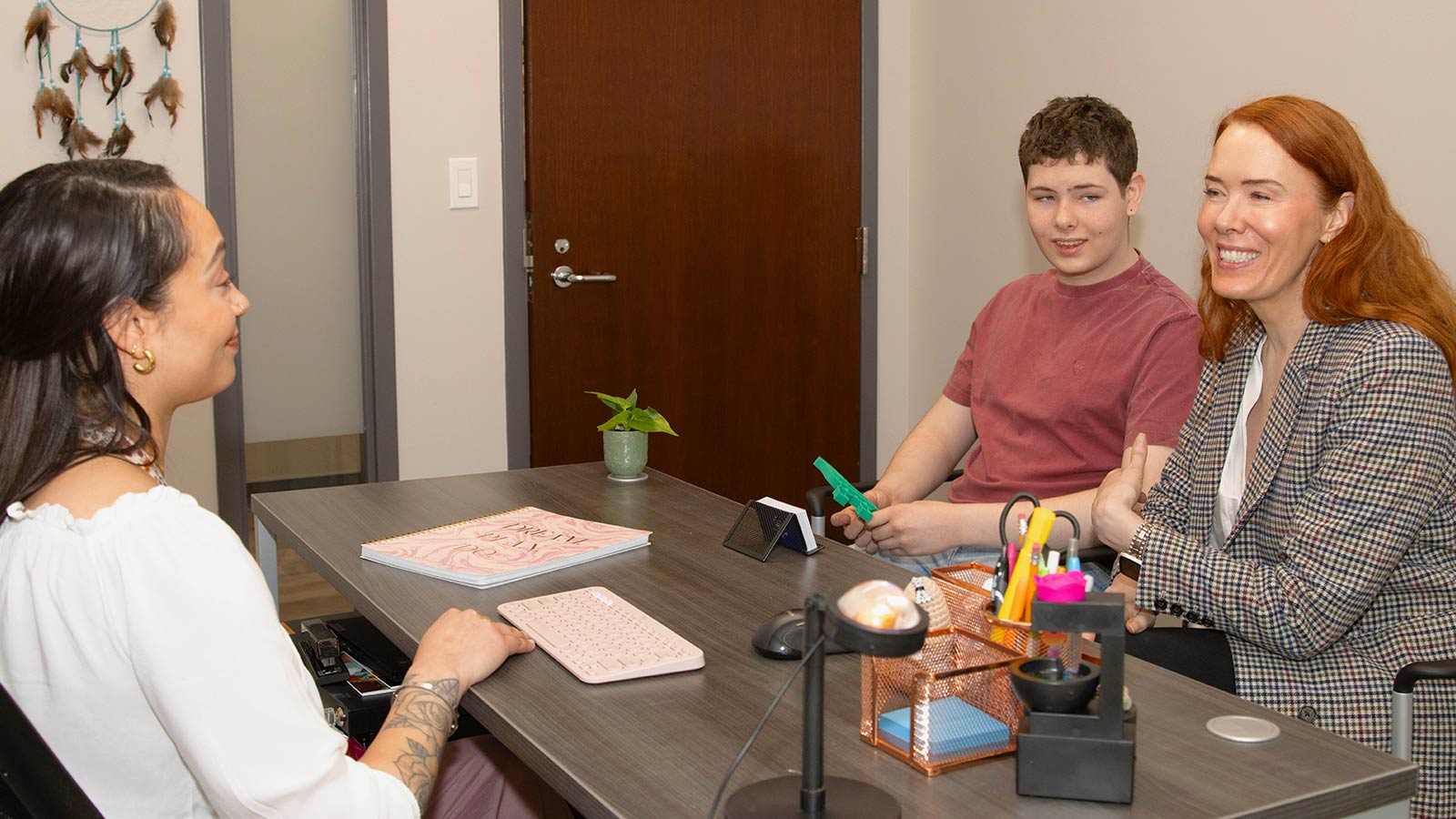 A female therapist is sitting at a desk across from a young man and his mother.