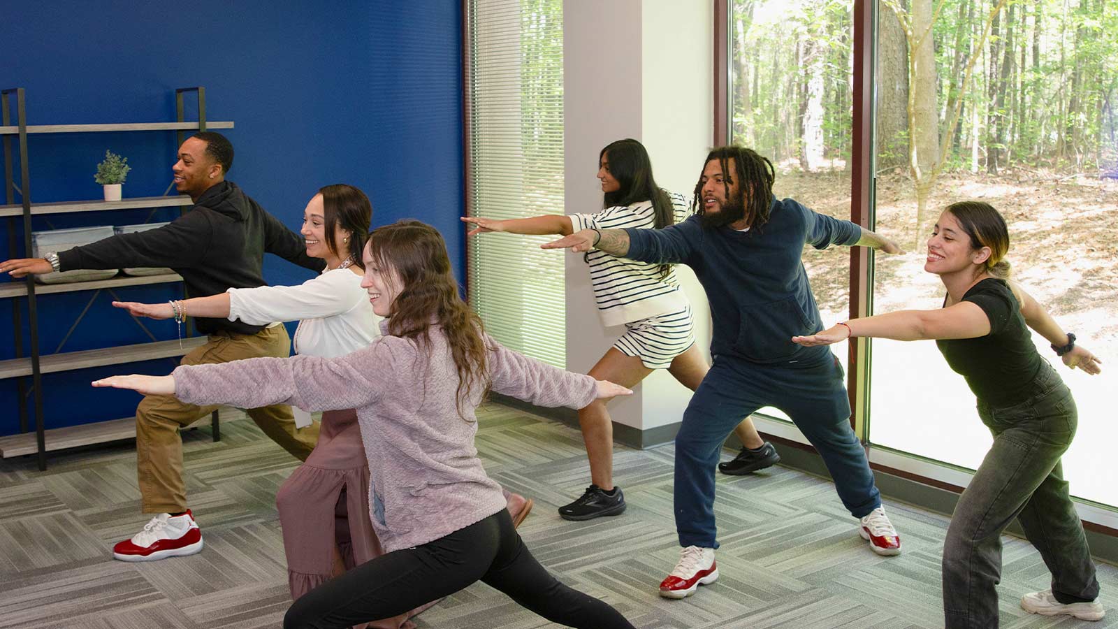A group of young adults practice yoga indoors in front of a large window.