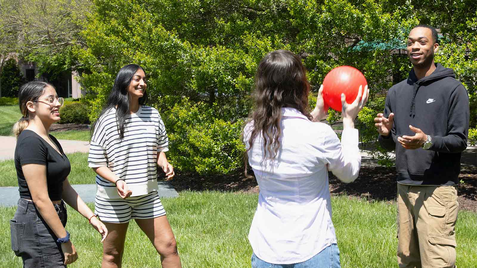 Four young adults play with a red ball outdoors on the grass.