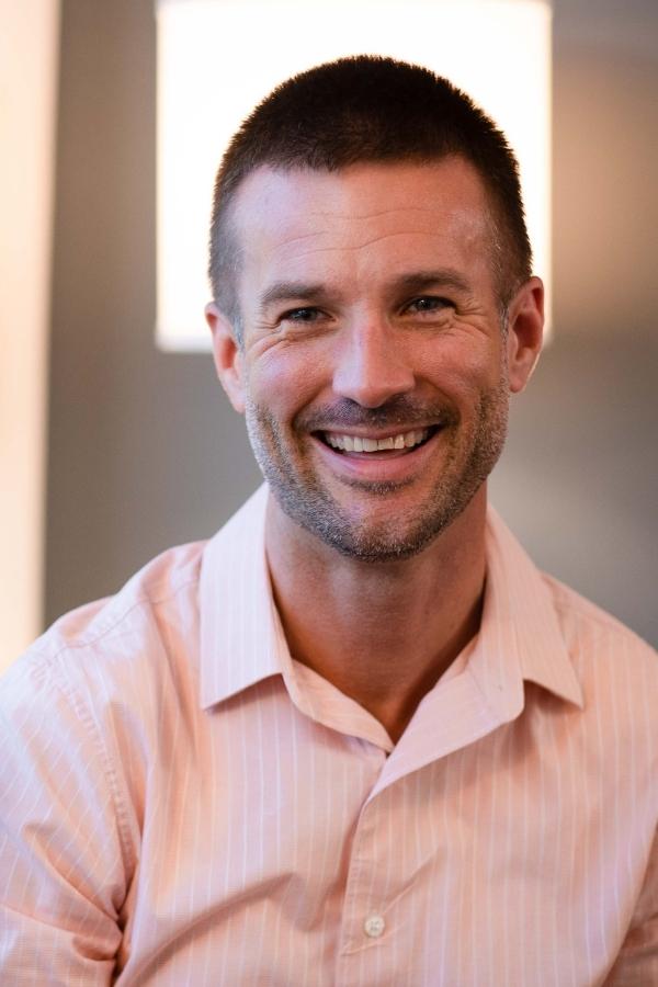 smiling man in pink shirt sitting in front of a window