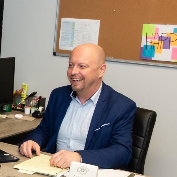 smiling man in blue suit sitting at desk with computer and papers