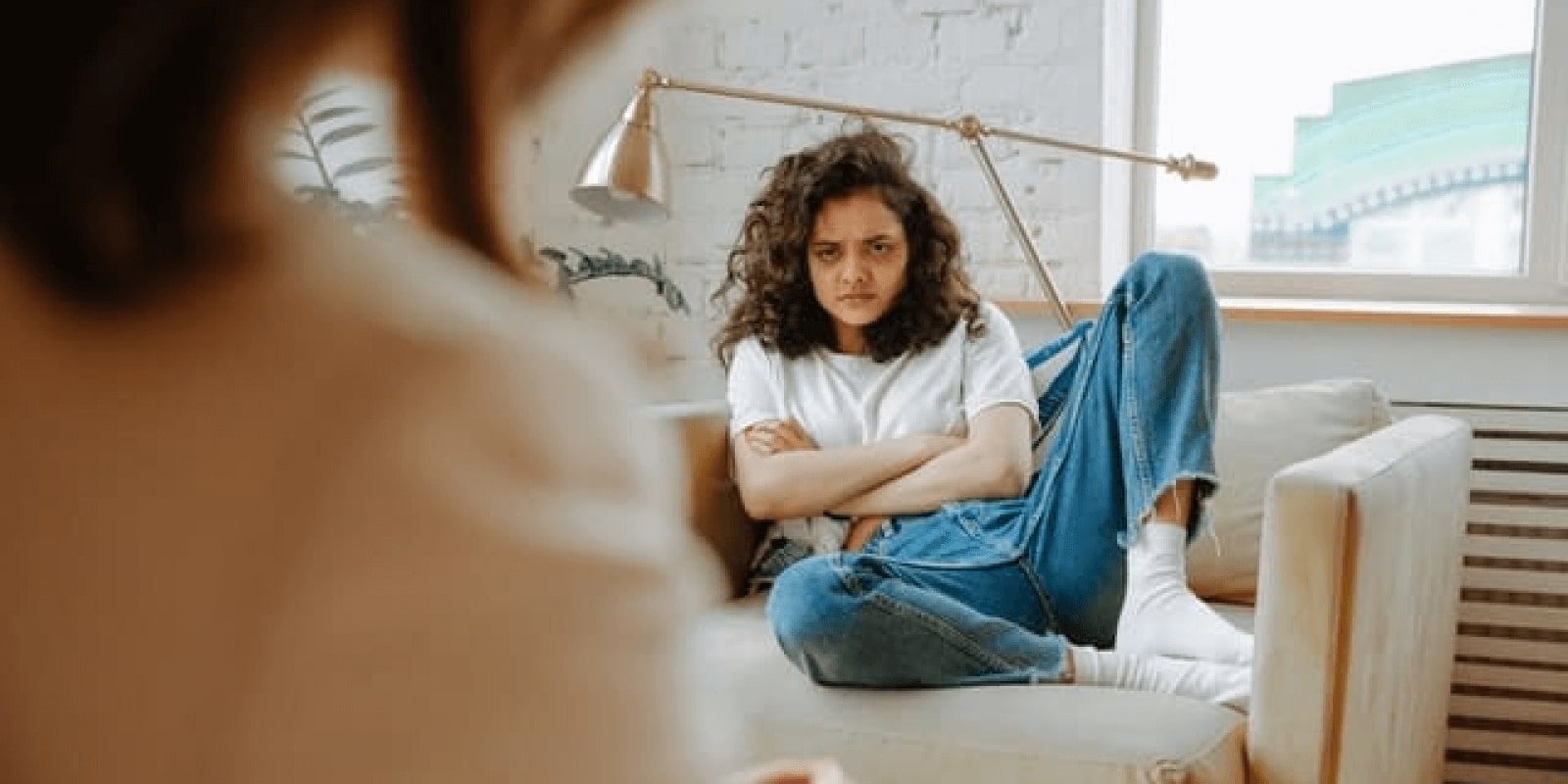 An irritated teen looking at her parent across a sofa with arms crossed