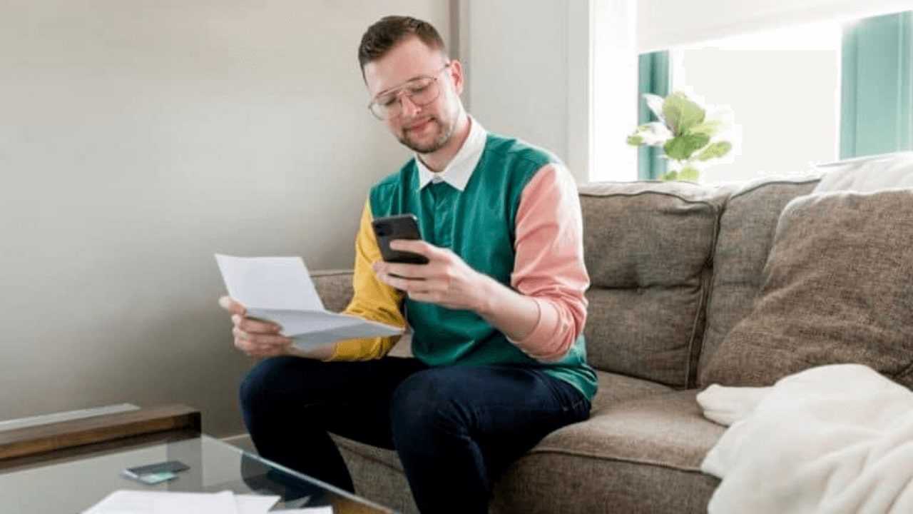 arafed man sitting on a couch reading a paper and looking at his cell phone