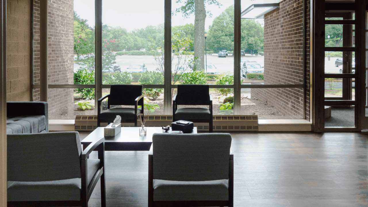 Sandstone Care's Reston Rehab Centers comfortable waiting area with modern chairs and a coffee table, set by a large window offering natural light and view of the parking lot and greenery outside.