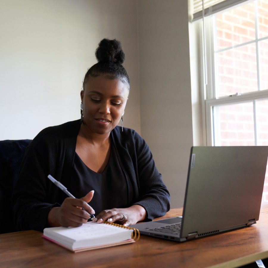 woman sitting at a desk writing in a notebook with a laptop in front of her