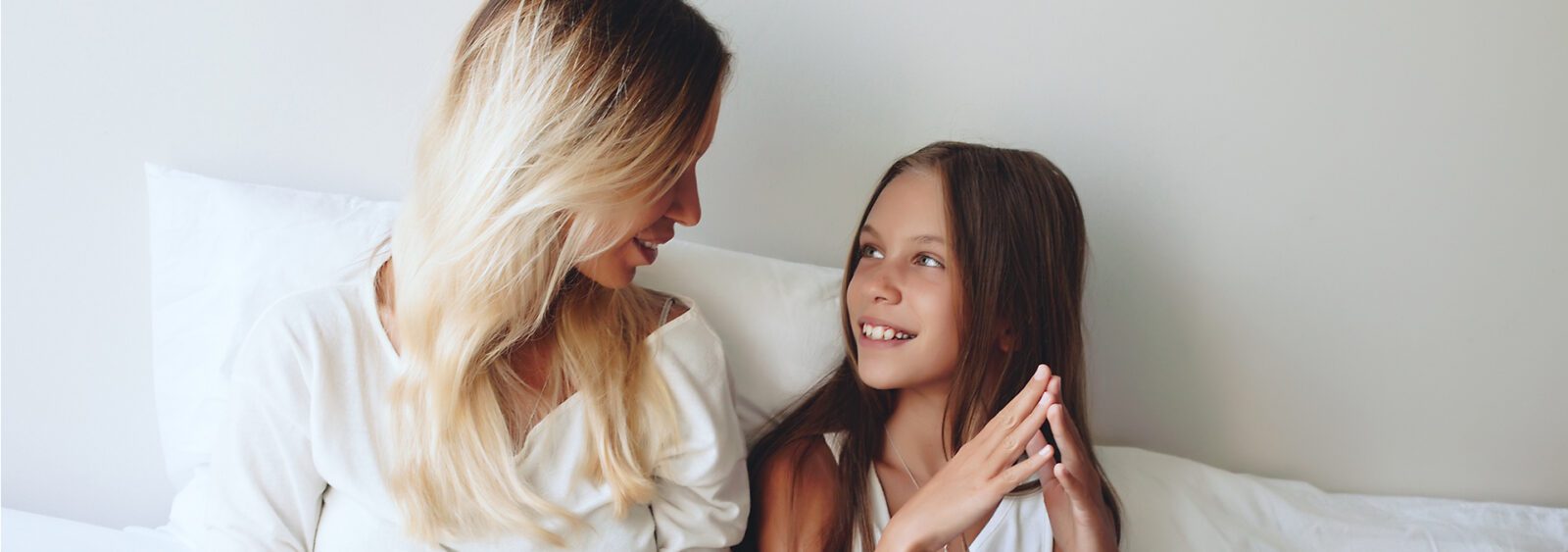 blonde woman and girl sitting on bed with white sheets and white sheets