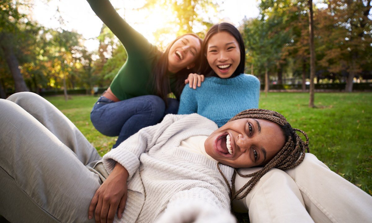 Three multiracial girls lying on the grass taking a selfie looking at the camera. Group of cheerful friends having a fun together, sharing happiness on social media. Concept of friendship and free