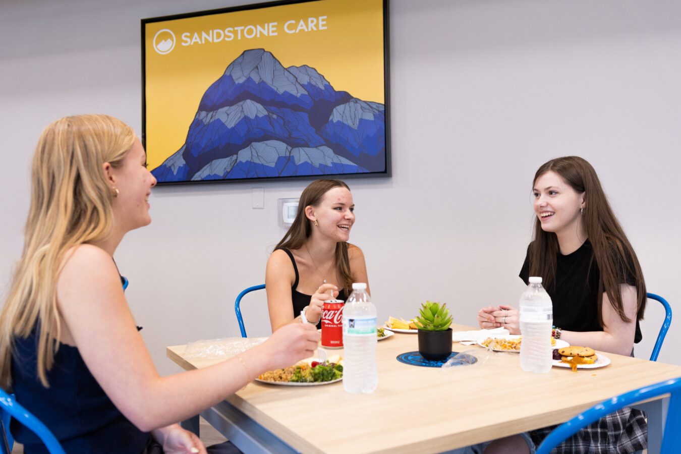 Three teenage girls eating and chatting in front of a sign that says Sandstone Care